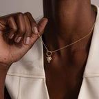 A person wearing a gold necklace with a Hummingbird Charm featuring a pear-cut diamond, holding it near their neck—a meaningful gift that sparkles with elegance.