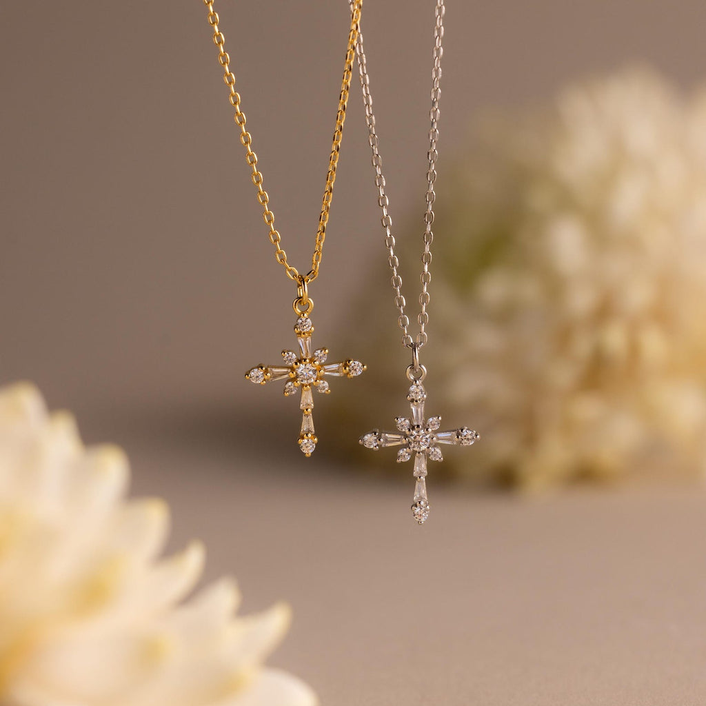 The Vintage Diamond Cross Necklace hangs against a soft beige background with blurred flowers, showcasing its sparkling gemstones in elegant gold and silver settings.