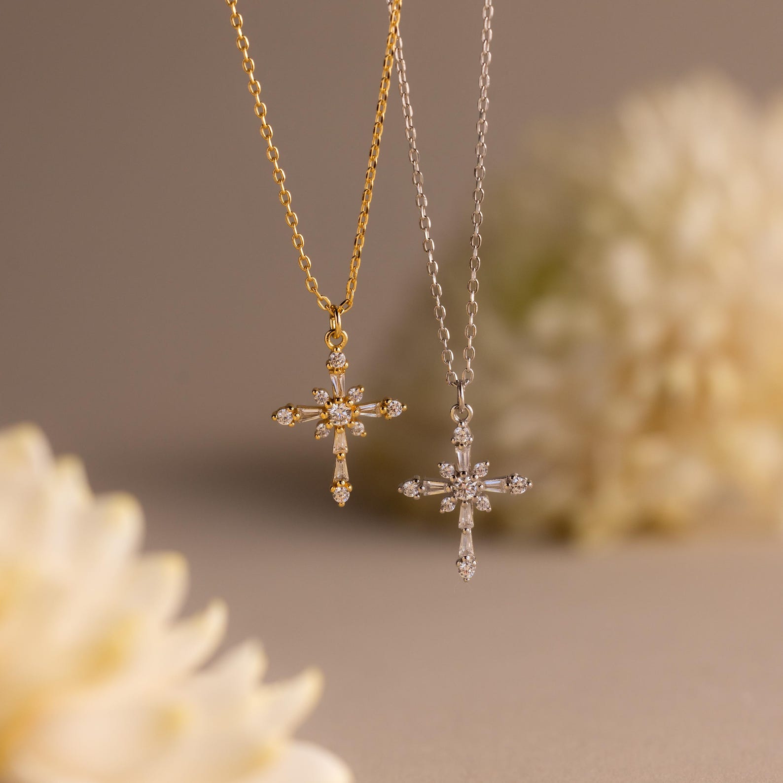The Vintage Diamond Cross Necklace hangs against a soft beige background with blurred flowers, showcasing its sparkling gemstones in elegant gold and silver settings.