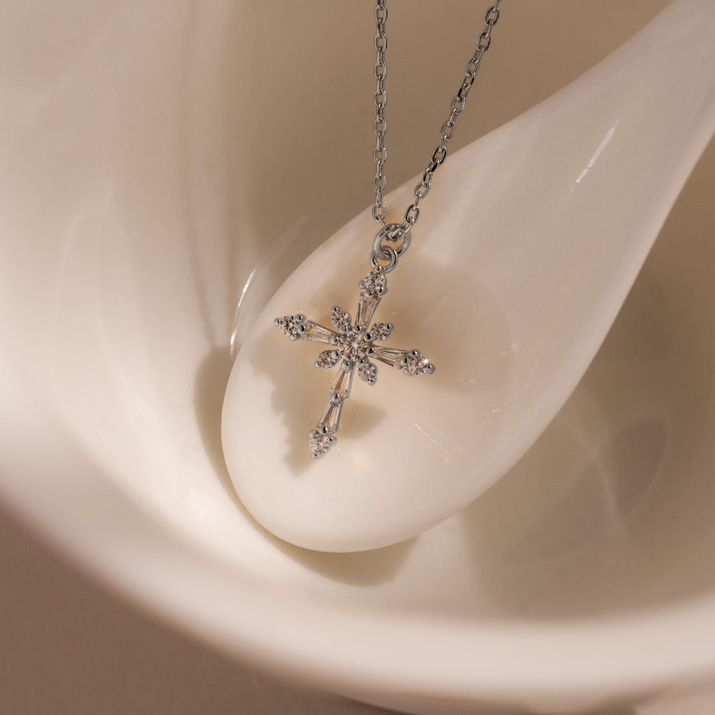 The Vintage Diamond Cross Necklace in Silver is elegantly displayed on a smooth white ceramic spoon nestled in a matching bowl.