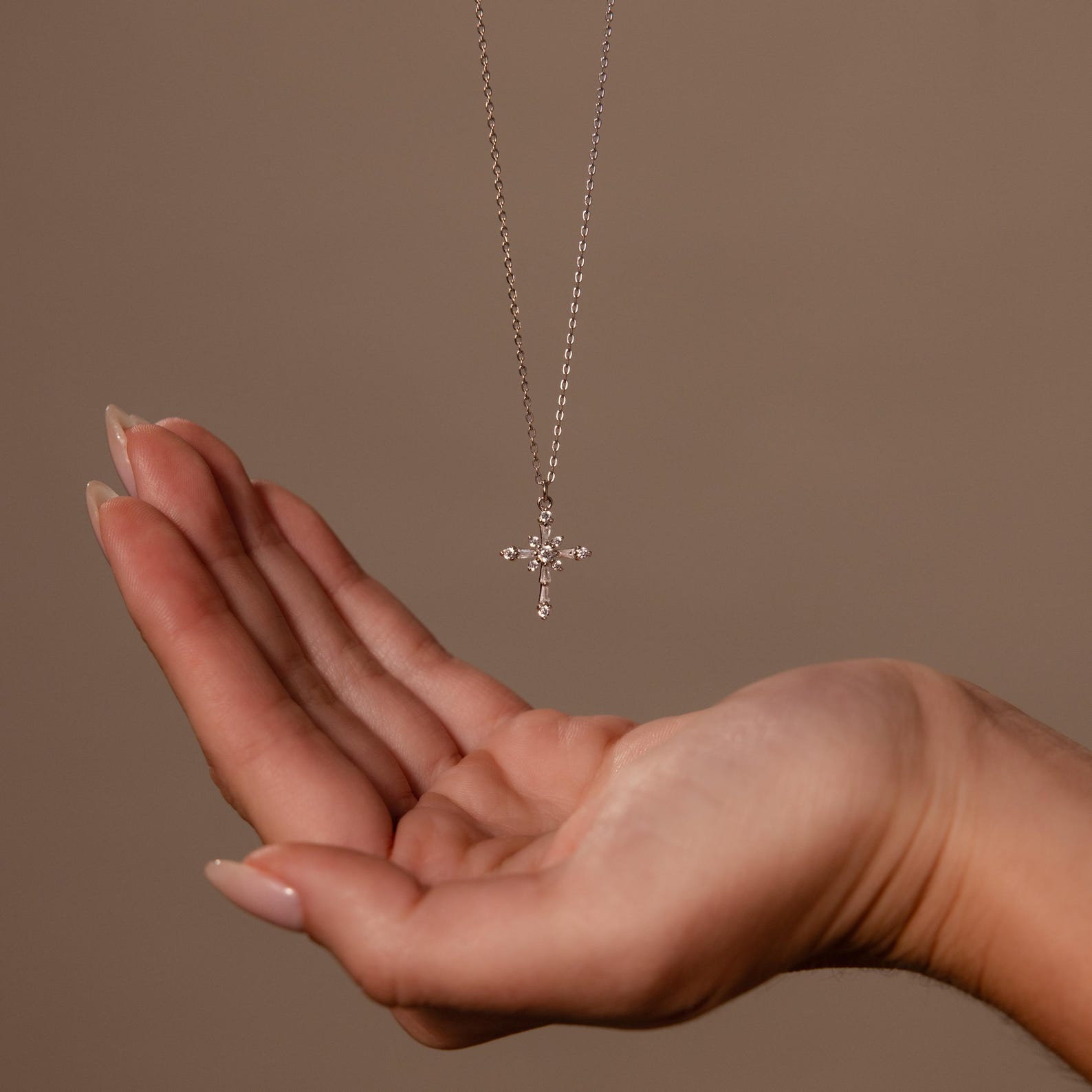 A hand reaches toward the Vintage Diamond Cross Necklace, its sparkling cross pendant gleaming against a plain beige background.