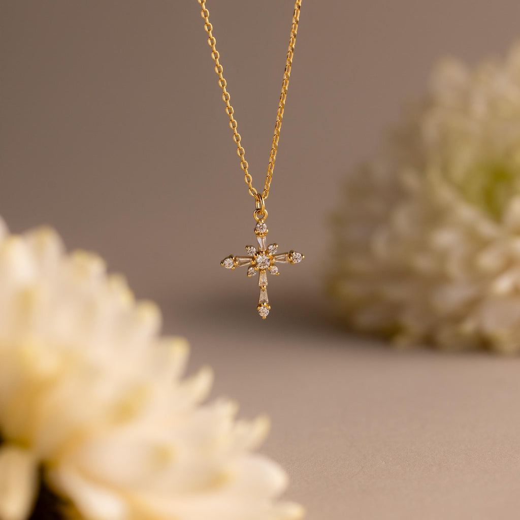 The Vintage Diamond Cross Necklace features a gold cross pendant with small diamonds, showcased against a soft-focus backdrop of white flowers.