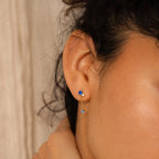 Close-up of an ear adorned with Sapphire Diamond Drop Earrings, showcasing a gold design with two blue gemstones—a sapphire stud and a dangling sapphire drop.