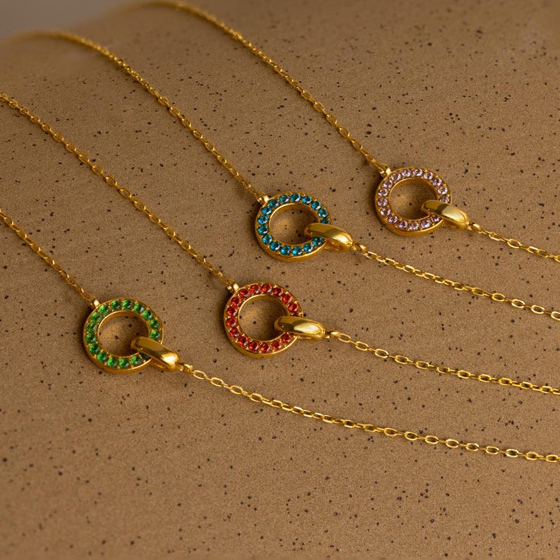 Four Interlocking Birthstone Necklaces with colorful gemstone circle pendants rest on a speckled beige surface—a timeless keepsake and a perfect gift for mom.