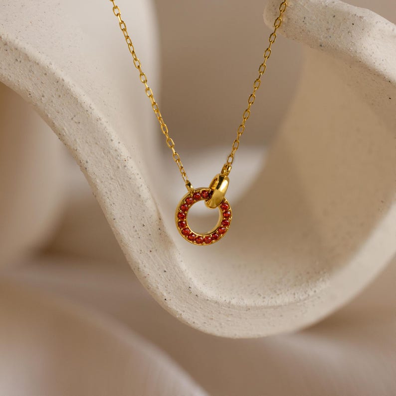 The Interlocking Birthstone Necklace features a gold chain and a small circular pendant set with a red gemstone, elegantly displayed on a beige surface—an ideal keepsake gift for mom.