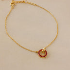 The Interlocking Birthstone Bracelet features a delicate gold chain with a red gemstone-studded charm, shown on a beige background—ideal for those who love customizable, personal birthstone jewelry.