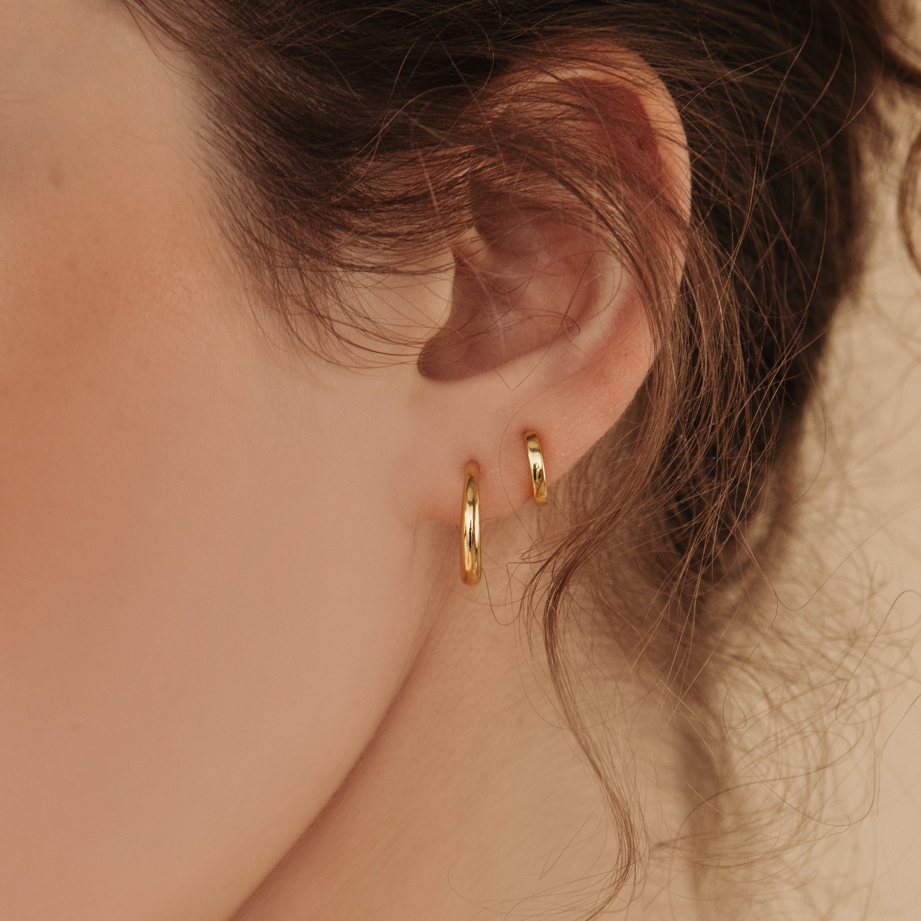 Close-up of a womans ear wearing the Editor Huggies in 18K Gold. Her brown hair is loosely pulled back, highlighting the minimal, classic style of these small gold earrings for an effortlessly elegant look.