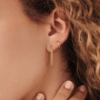 A womans ear is styled with the Editor Huggies in 18K Gold on her upper lobe and a chain dangle below. Her curly hair and graceful hand highlight the timeless elegance of this classic jewelry arrangement.