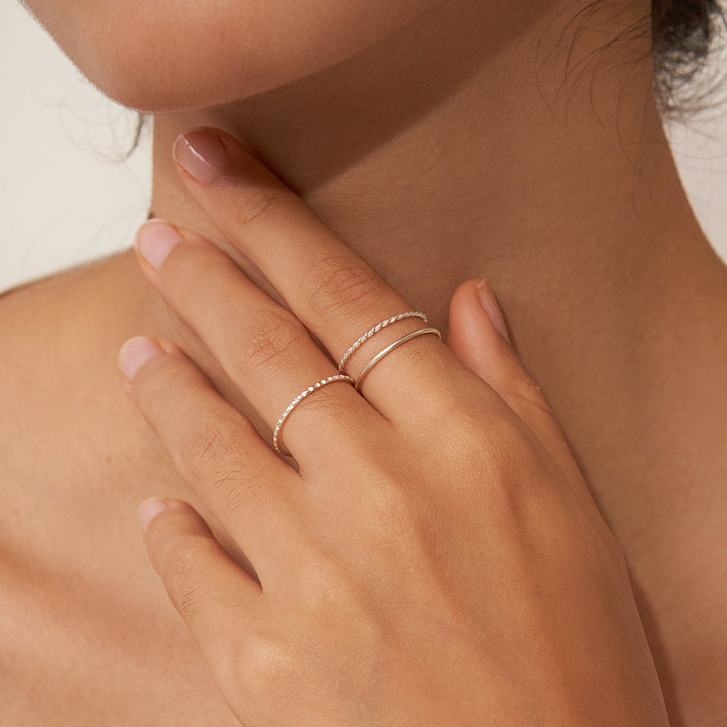 A hand with nude nails, adorned with the Thin Stacking Rings Set in Sterling Silver, gently touches a woman's neck.