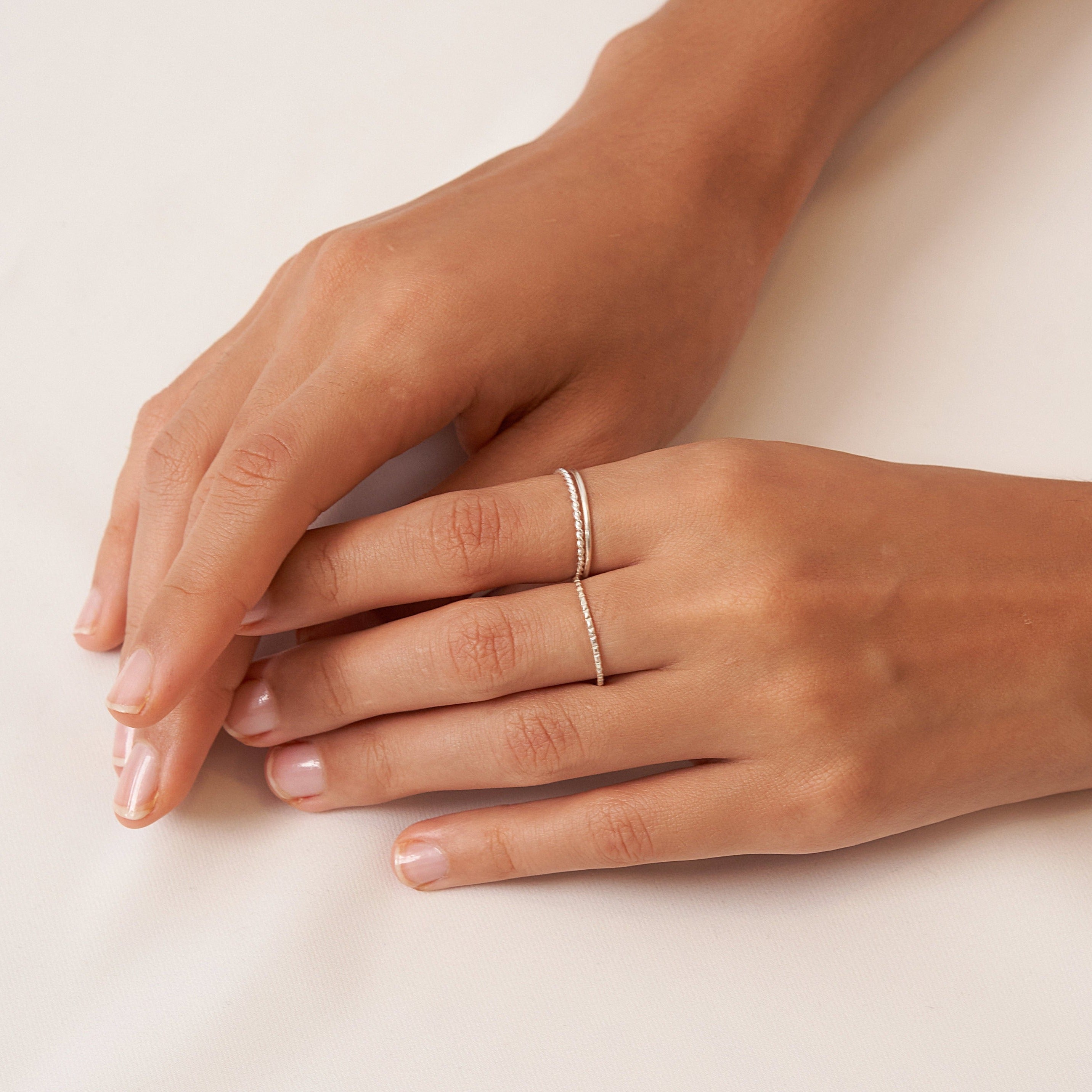 Two hands on a white surface, one adorned with the Thin Stacking Rings Set in Sterling Silver—an elegant and stylish Christmas gift choice.