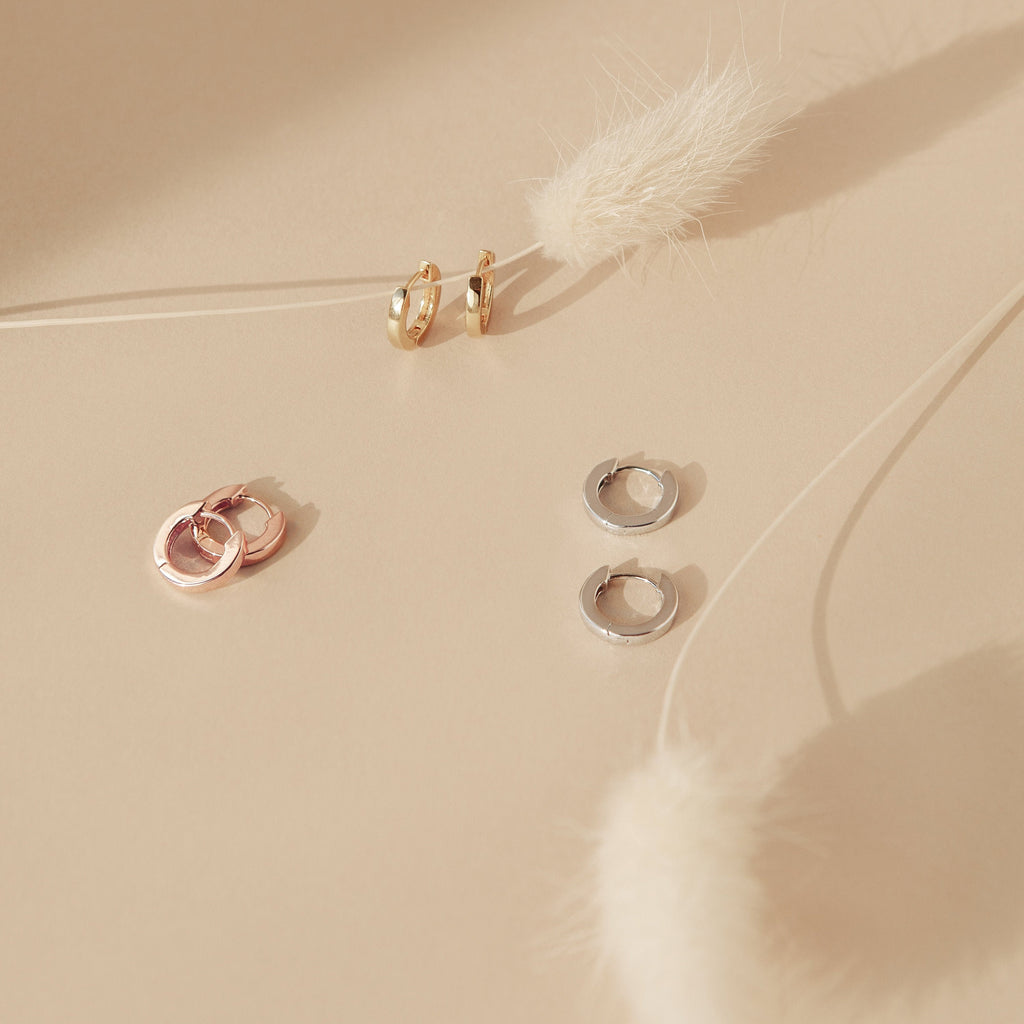 Three pairs of Editor Huggies—in gold, rose gold, and silver—are displayed on a beige surface with delicate white dried flowers for a minimal, elegant look.