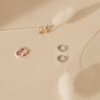 The Editor Huggies in 18K Gold are minimal earrings, shown alongside rose gold and silver pairs on a beige surface with white dried florals—ideal for those who love classic jewelry.