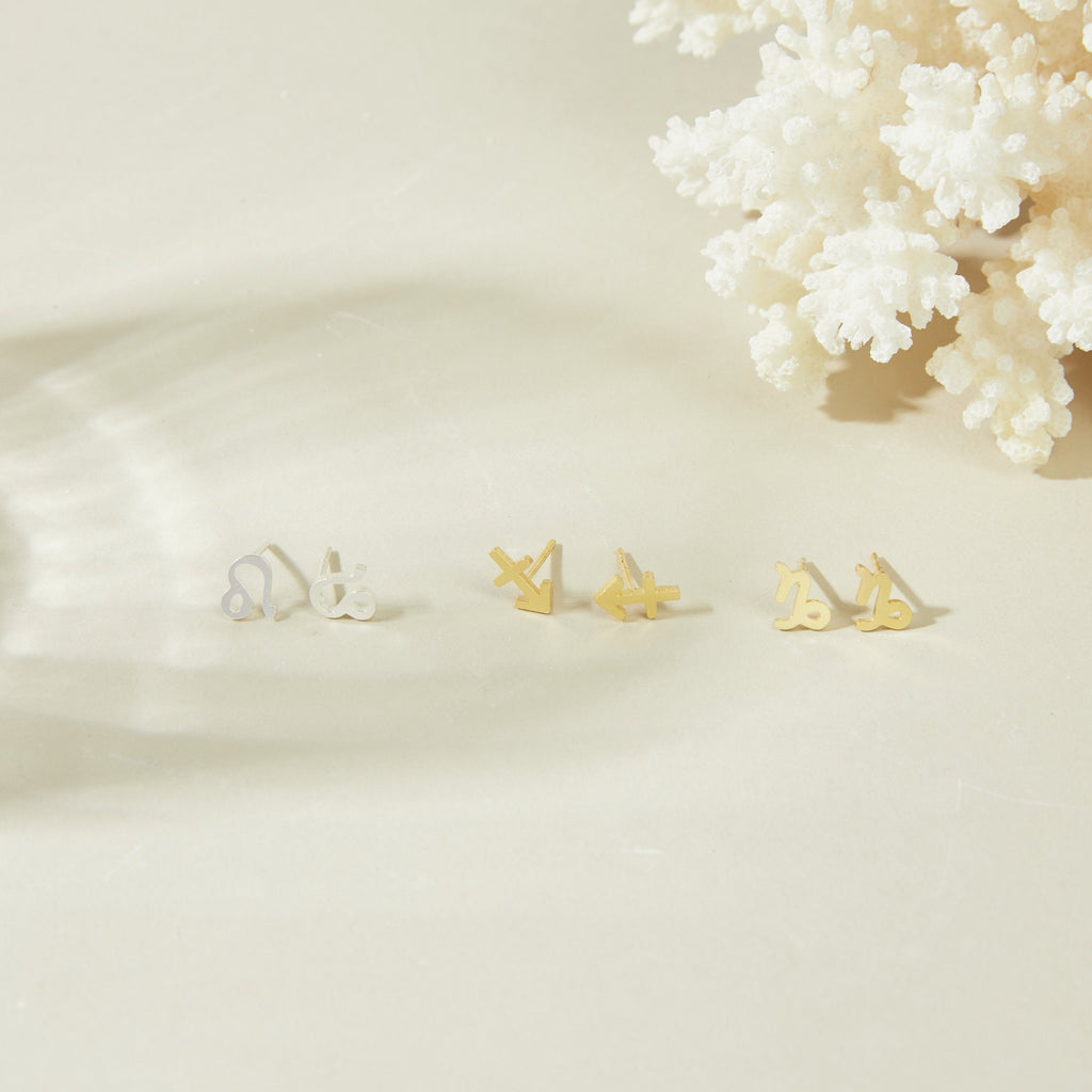 A set of three pairs of minimalist stud earrings, including Tiny Zodiac Studs in gold and silver, is displayed on a beige surface beside white coral.