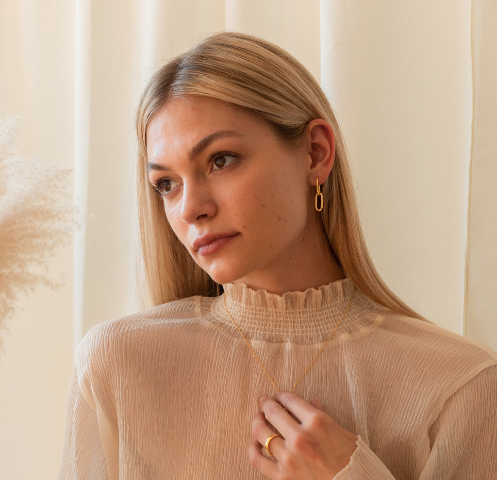 A woman with long blonde hair wears gold Linked Earrings Set from her jewelry collection and a cream ruffled blouse as she gazes thoughtfully to the side.