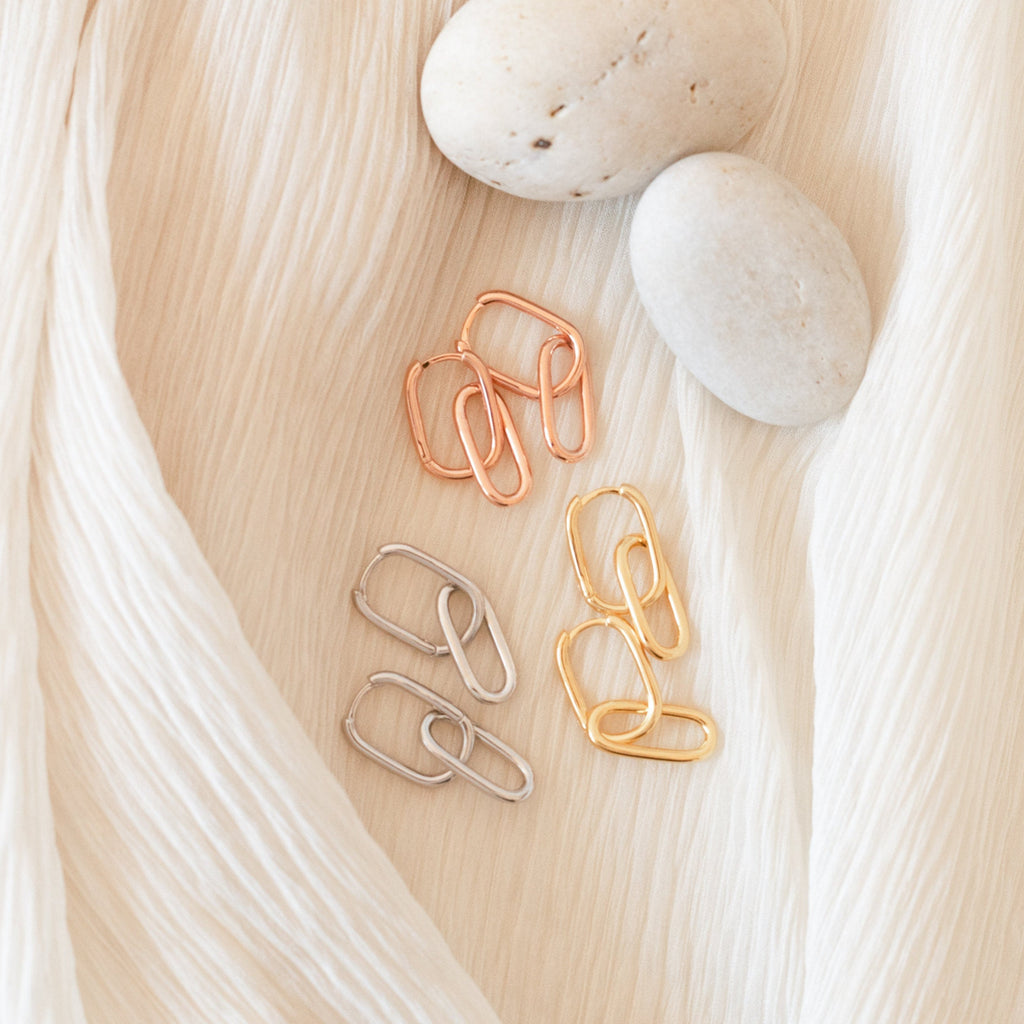 Three pairs of Amanda Link Earrings in gold, rose gold, and silver rest beside smooth stones on cream fabric—a refined take on classic minimal knot earrings.