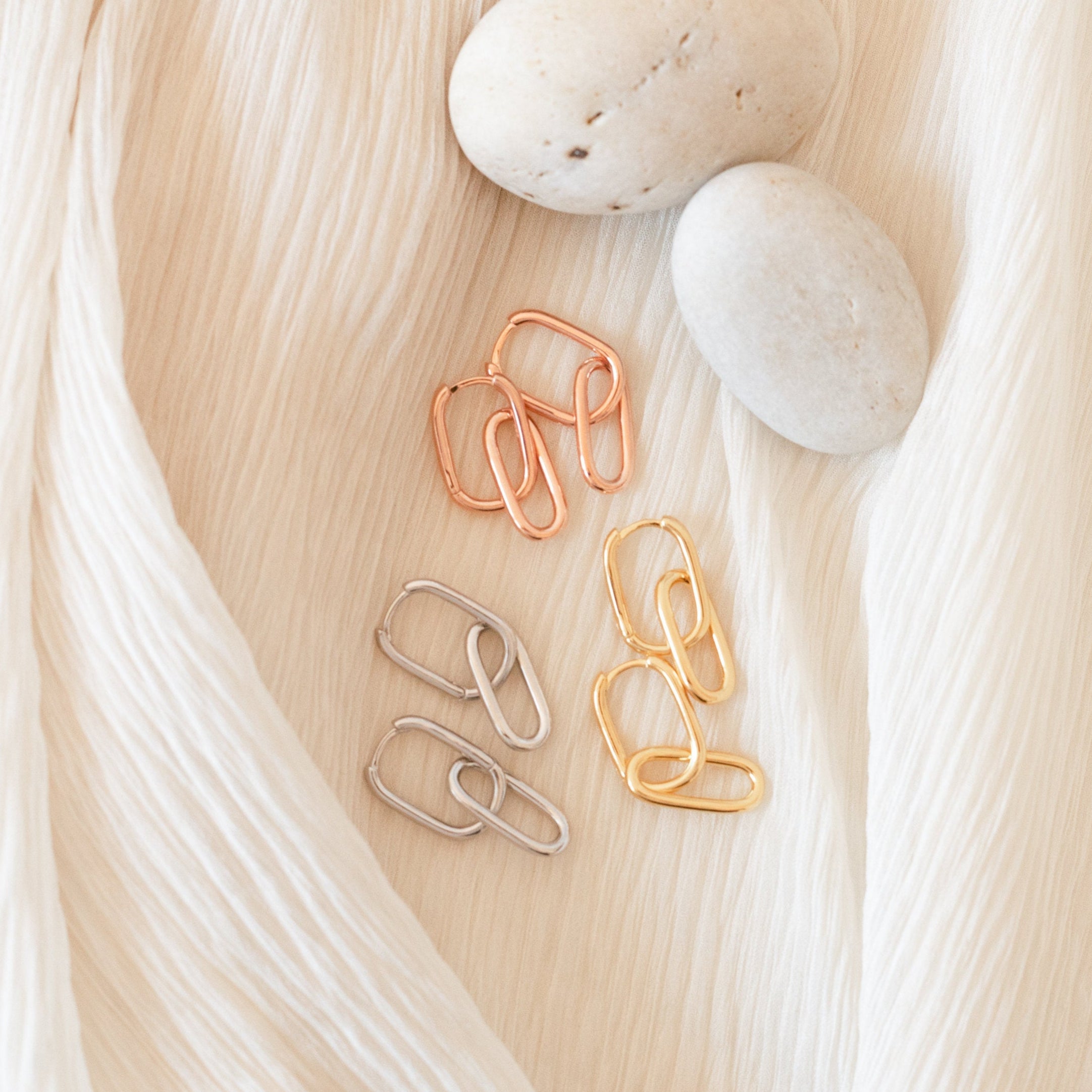 Three pairs of Amanda Link Earrings in gold, rose gold, and silver rest beside smooth stones on cream fabric—a refined take on classic minimal knot earrings.