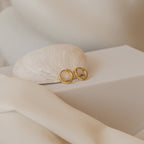 The Editor II Huggies—small gold hoop earrings—are shown on a white seashell atop a white box with soft fabric folds, offering a timeless, classic style perfect for everyday wear.