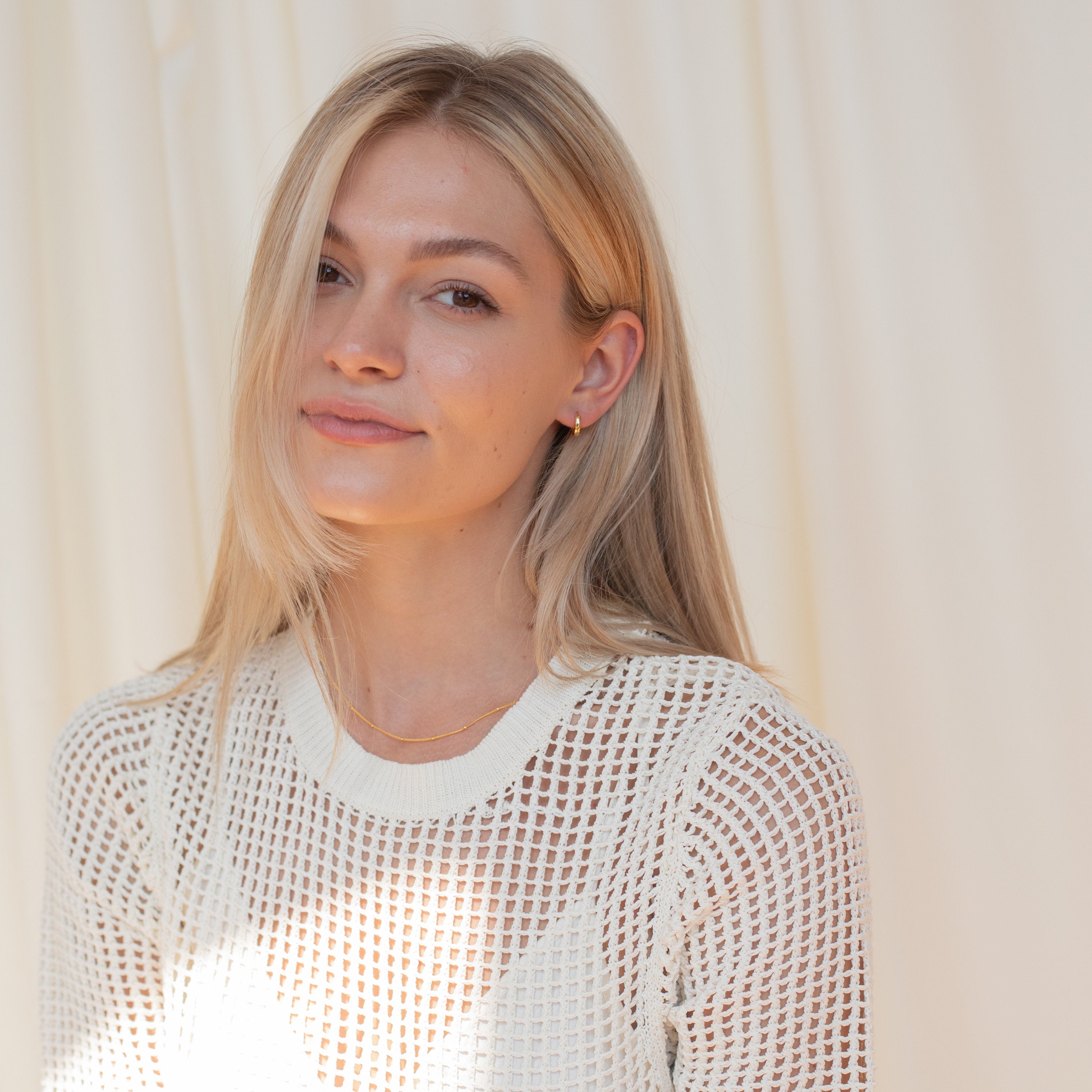 A woman with straight blonde hair in a white mesh top smiles softly against a light background, her look completed with the Editor II Huggies.