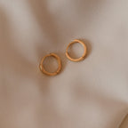 Two Editor II Huggies in sterling silver rest on a beige fabric background, perfect for everyday wear or adding subtle elegance—these earrings are a timeless essential.