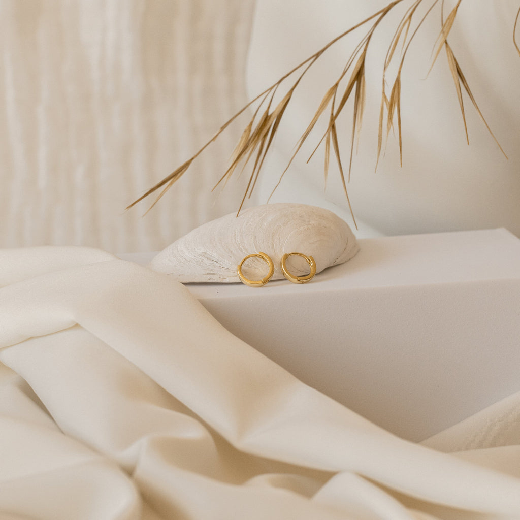 The Editor II Huggies in 18K Gold rest on a seashell, with beige fabric and dried grass behind them—perfect everyday earrings for adding effortless style to any outfit.