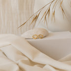 The Editor II Huggies in 18K Gold rest on a seashell, with beige fabric and dried grass behind them—perfect everyday earrings for adding effortless style to any outfit.