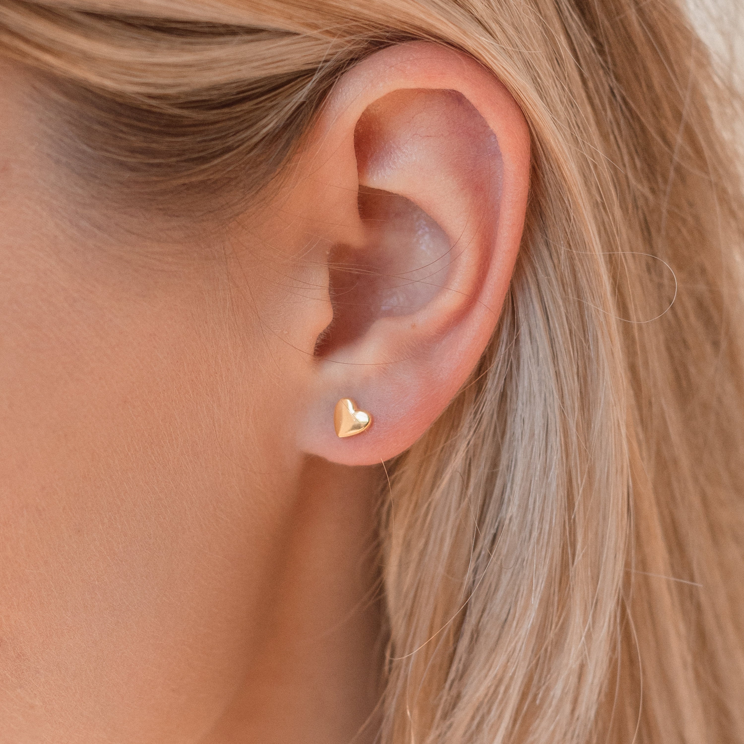Close-up of a model wearing a gold heart-shaped stud earring with blonde hair.