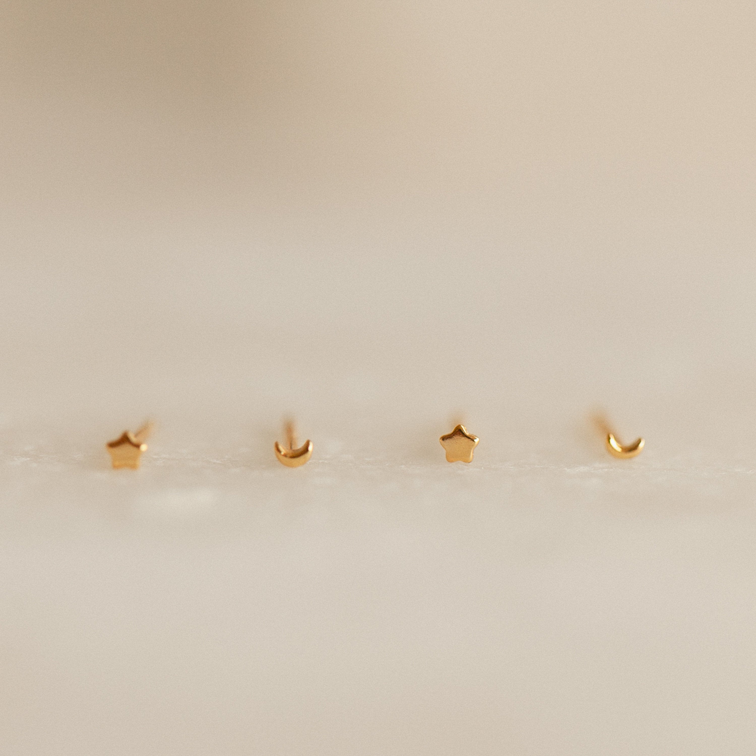 Four gold earrings shaped like stars and moons on a beige background