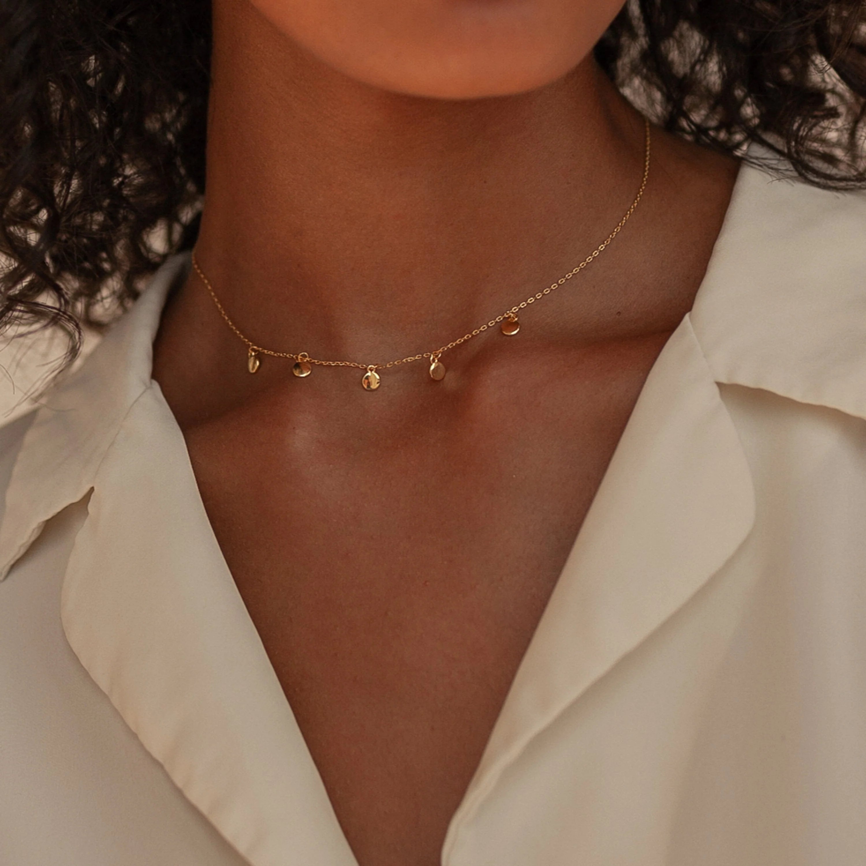A woman in a cream collared shirt showcases minimalist style, layering a gold charm necklace with the Onyx Necklace Set.