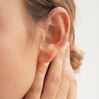 Close-up of a person's ear adorned with Tiny Moon & Star Studs in 18K Gold, alongside two small gold hoop earrings, as a hand gently touches the ear.