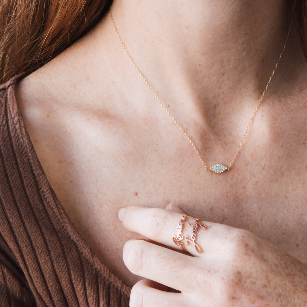 A woman wearing the Opal Eye Necklace—a delicate gold necklace with an opal eye pendant—and a gold ring, gently touching her collarbone.