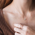 A woman wearing the Opal Eye Necklace—a delicate gold necklace with an opal eye pendant—and a gold ring, gently touching her collarbone.