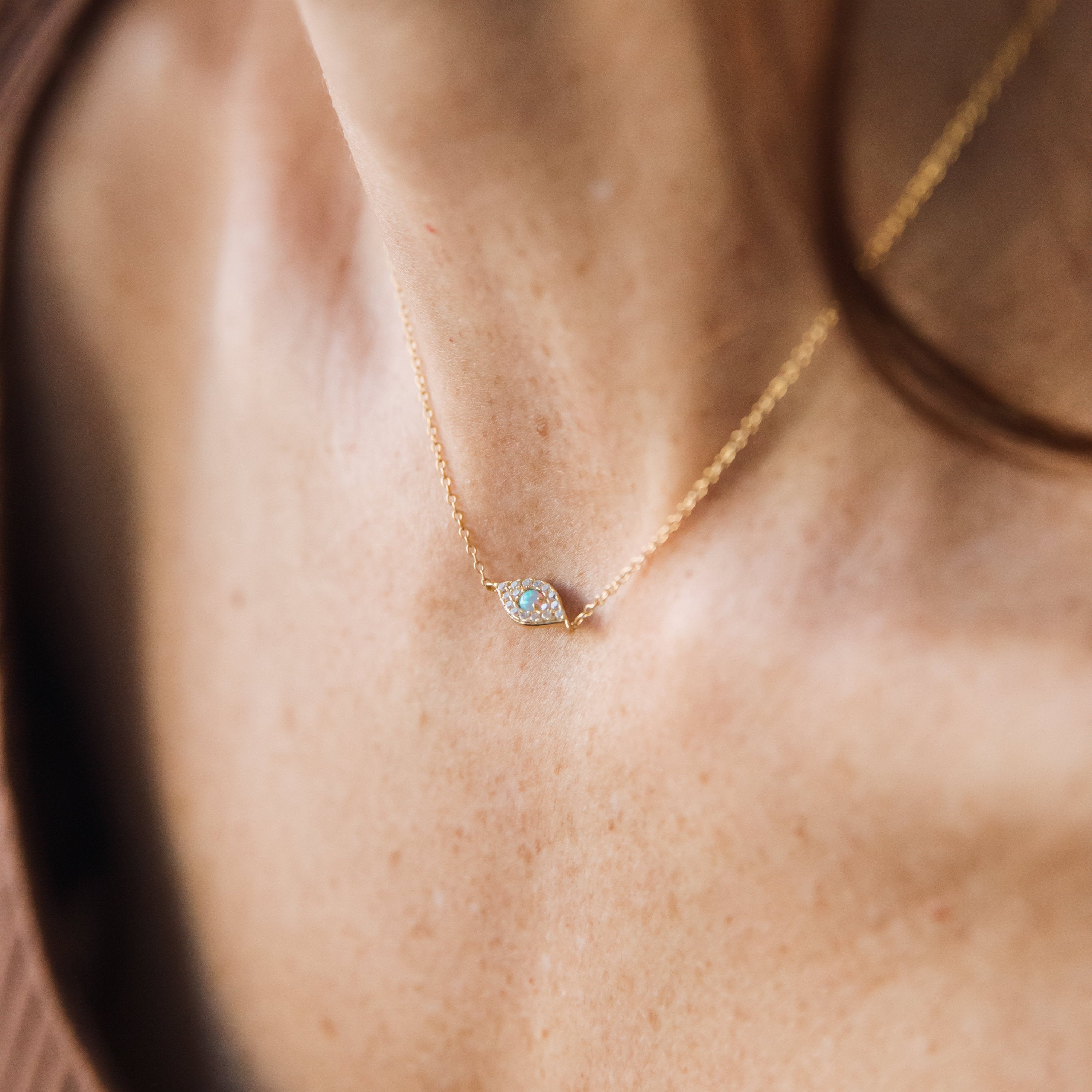 The Opal Eye Necklace, featuring a delicate gold chain and small opal eye pendant, rests gracefully on a womans neck.