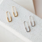 The Safety Pin Earrings Set includes two pairs of minimalist earrings—one gold, one silver—styled like paperclips and displayed on a textured white surface for a subtle yet chic statement.