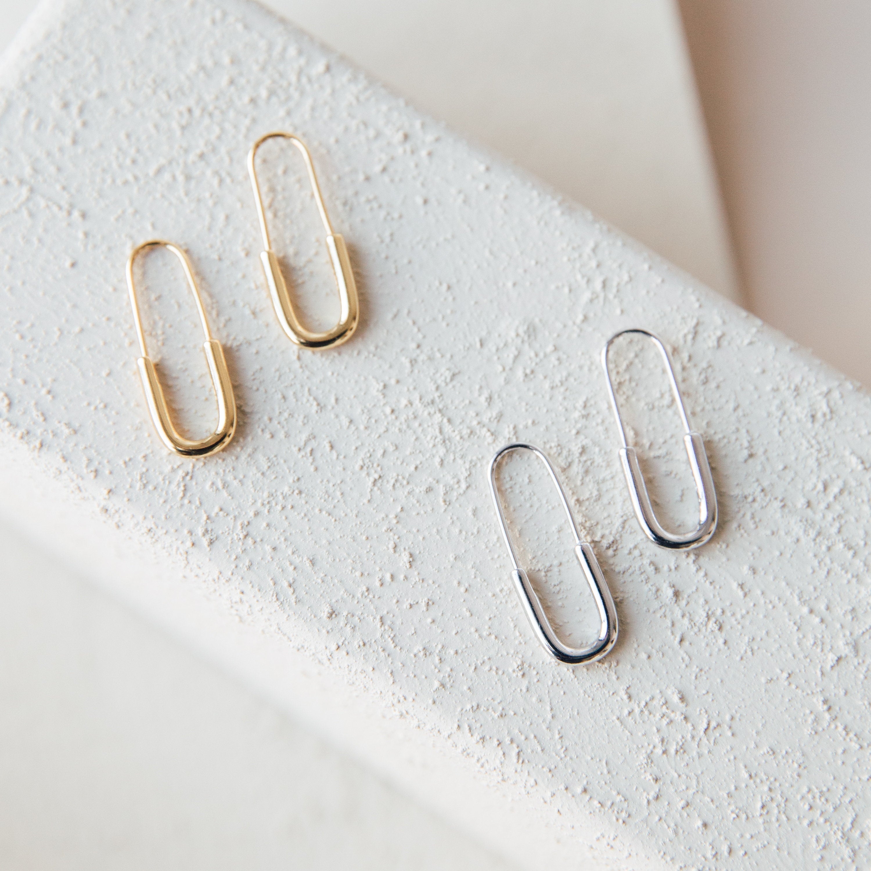 The Safety Pin Earrings Set includes two pairs of minimalist earrings—one gold, one silver—styled like paperclips and displayed on a textured white surface for a subtle yet chic statement.