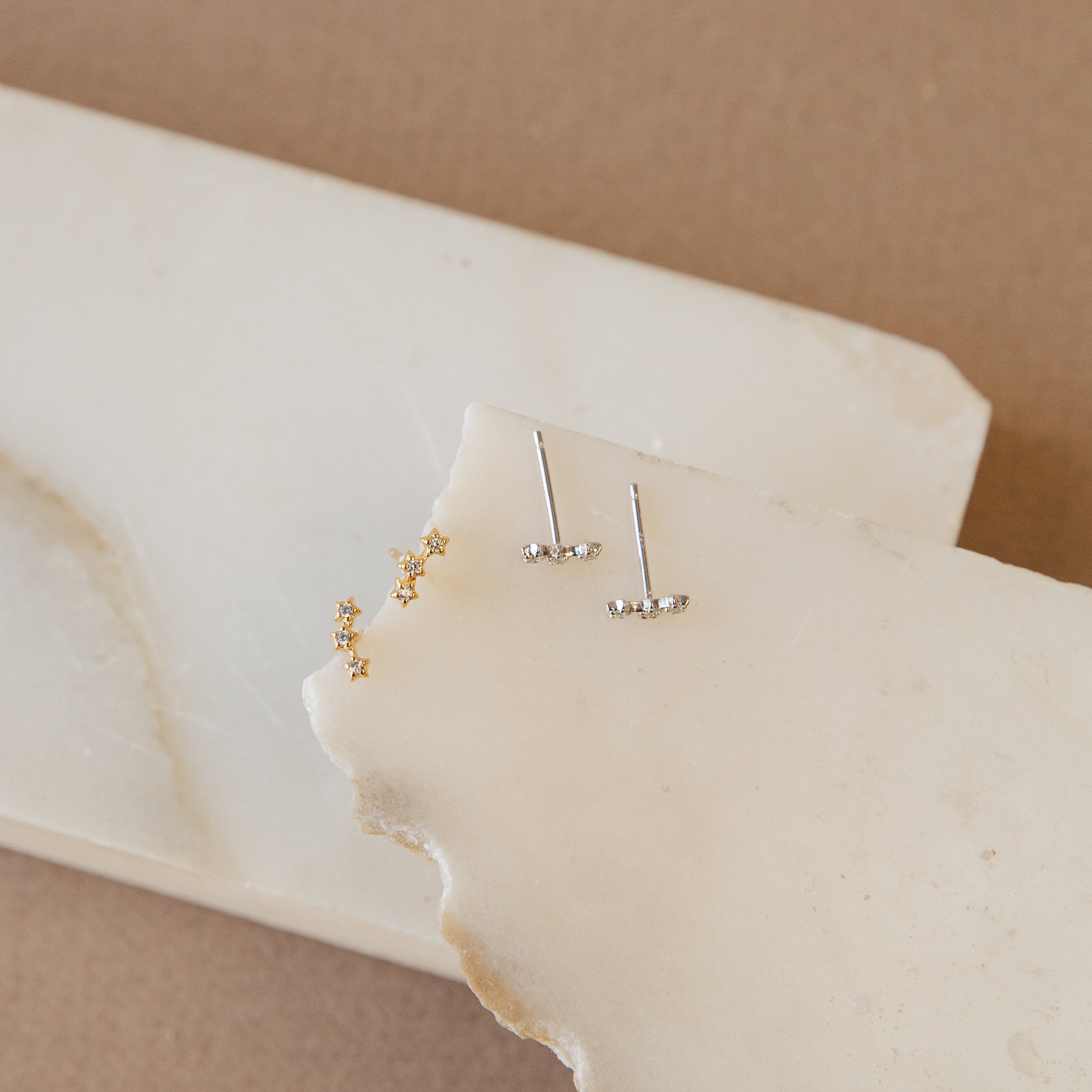 Two pairs of gold and silver stud earrings on a white stone surface with a beige background