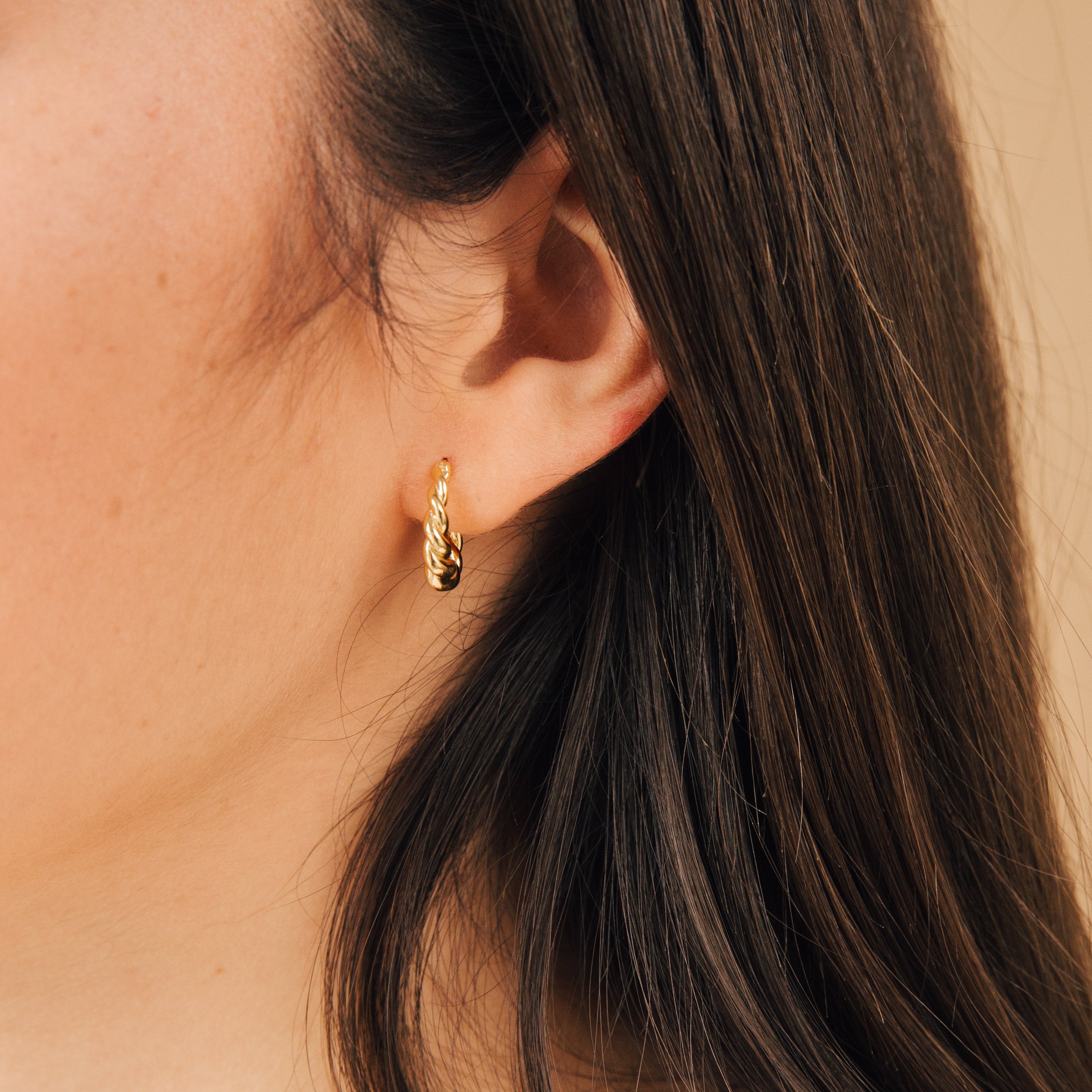 Close-up of a woman’s ear adorned with Naomi Twisted Hoops in 18K Gold, her long brown hair cascading beside the delicate earrings.