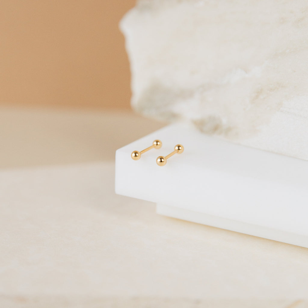 Nora Duo Studs in 18K Gold—small, classic ball stud earrings displayed on white geometric surfaces with a beige background—perfect for everyday wear.