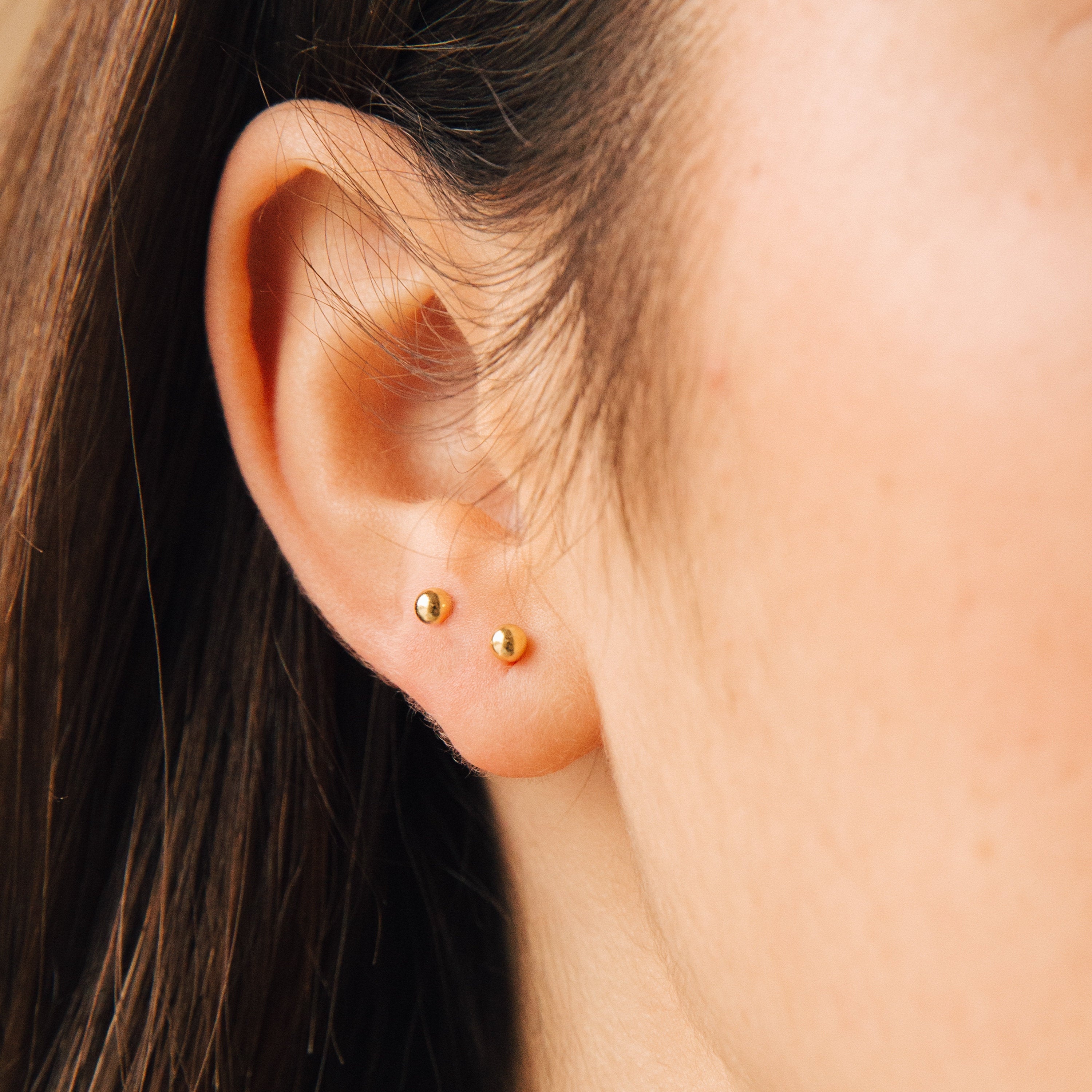 Close-up of an ear with two Nora Duo Studs, small gold ball earrings in the lower lobe—classic everyday style—paired with straight brown hair.