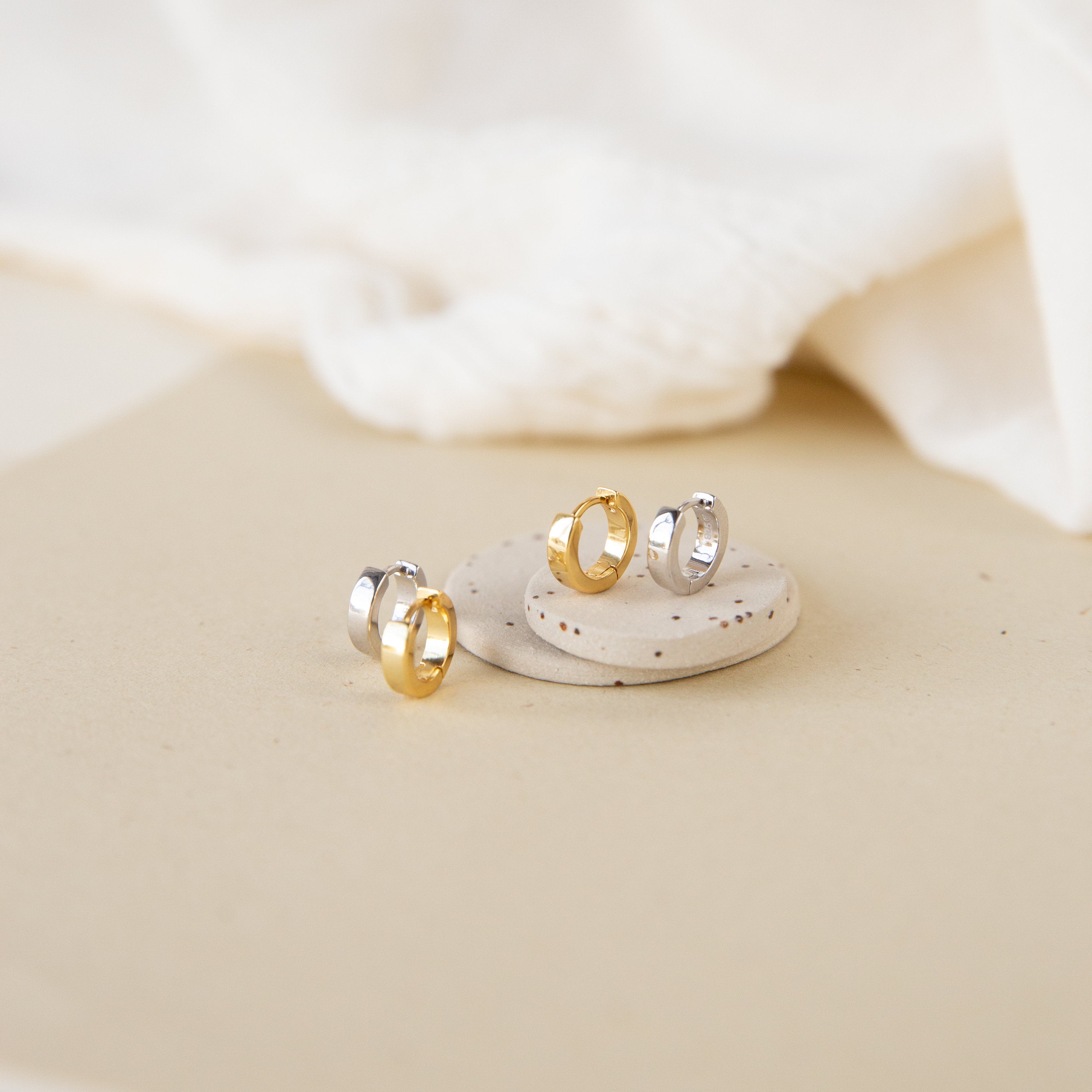 A pair of Meg Huggies in Sterling Silver displayed on a beige stone with soft fabric in the background.