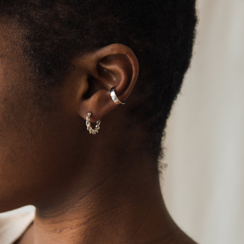 Close-up of a person's ear adorned with Naomi Twisted Hoops, a silver hoop earring, and a silver ear cuff, all set against a neutral background.