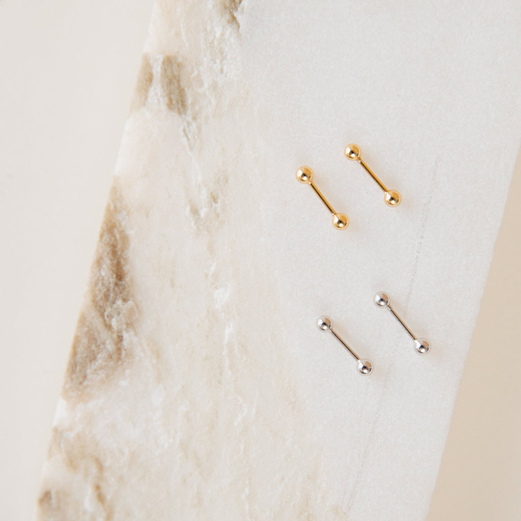 The Nora Duo Studs, featuring two gold and two silver barbell earrings, are classic accessories displayed on a white and beige marbled surface.