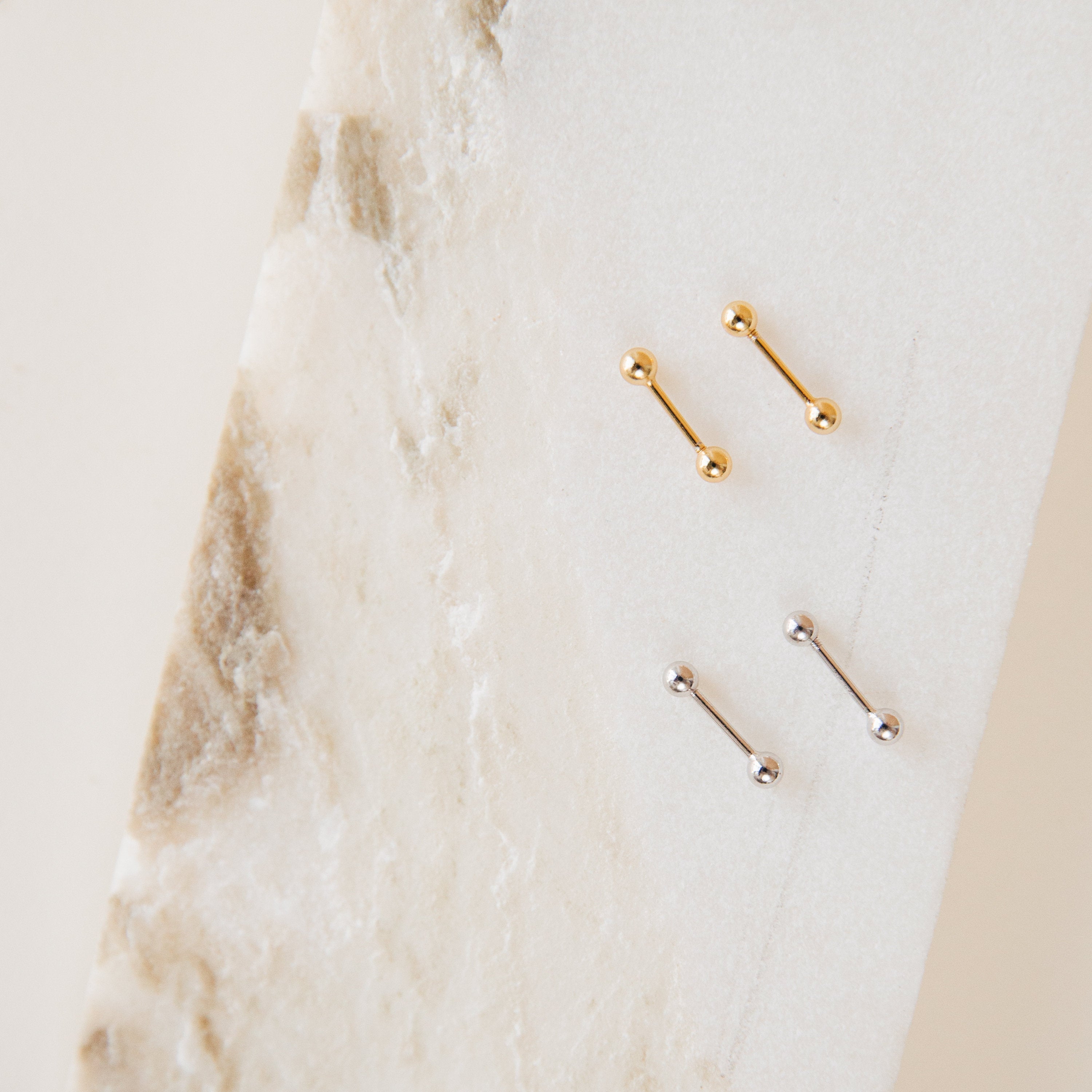 The Nora Duo Studs, featuring two gold and two silver barbell earrings, are classic accessories displayed on a white and beige marbled surface.