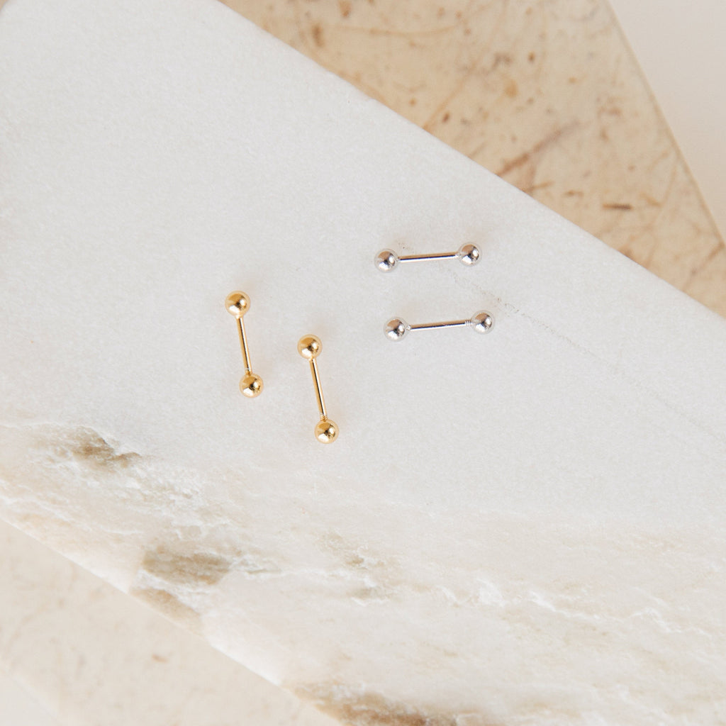 The Nora Duo Studs feature two pairs of classic barbell earrings, one in gold and one in silver, displayed on a white and beige marble surface—perfect for everyday wear.