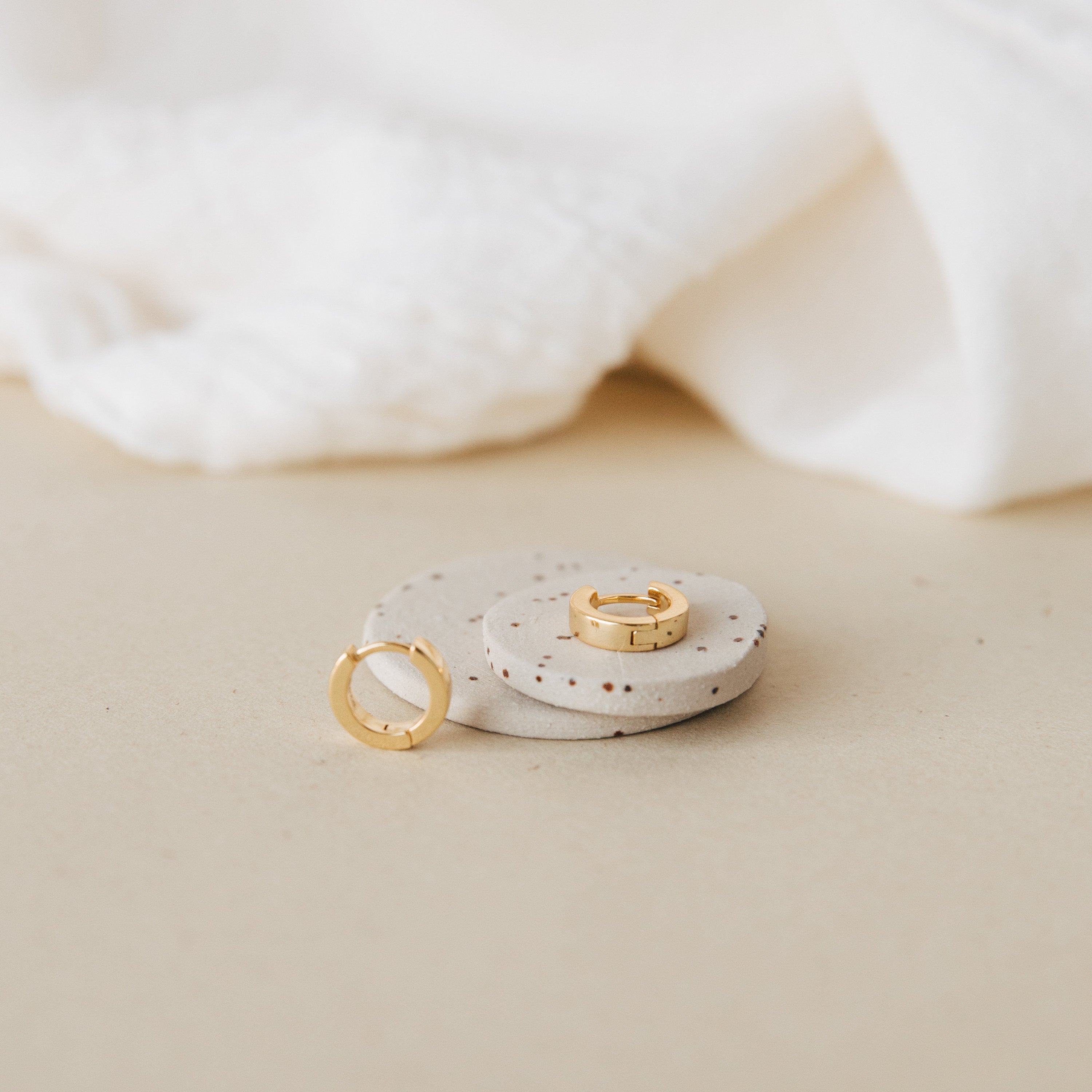 The Meg Huggies in 18K Gold are displayed on round speckled stones, set against a backdrop of soft white fabric.
