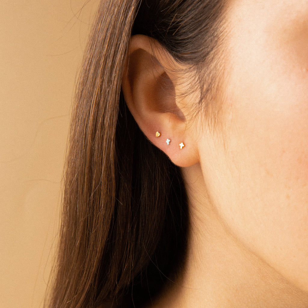 Close-up of a woman's ear adorned with three dainty earrings, including Tiny Cross Studs in 18K Gold, against a neutral background.