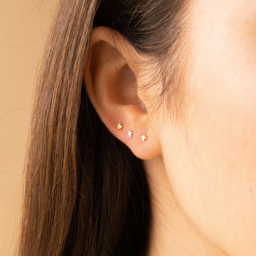 Close-up of an ear adorned with three dainty earrings: a heart, a star, and Tiny Cross Studs in Sterling Silver.