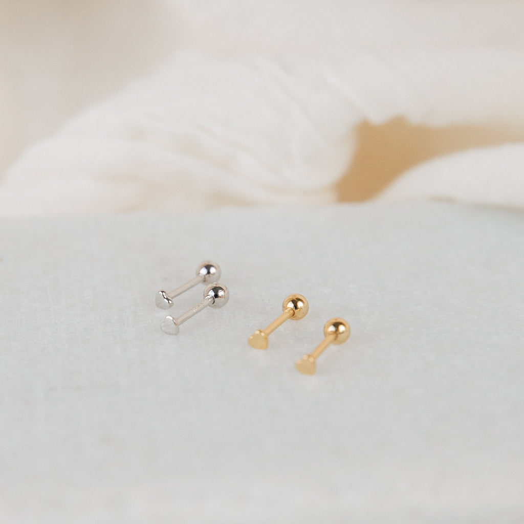 Two pairs of Tiny Heart Studs—one silver, one gold—are shown on a light textured surface. These earrings are perfect for everyday wear and make a thoughtful gift for someone special.