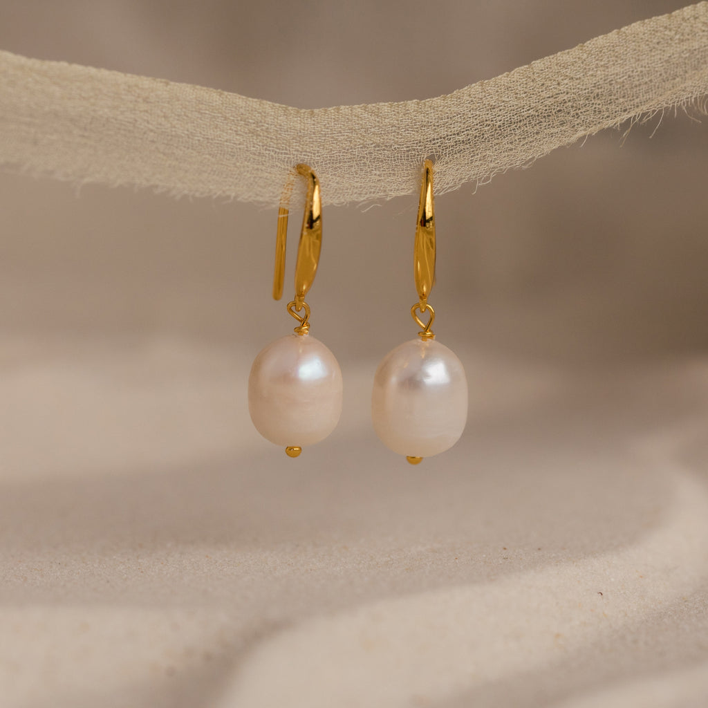 A close-up view of the Dangling Pearl Drop Earrings delicately hanging from a textured beige ribbon. Each earring features a luminous, oval-shaped freshwater pearl suspended from a sleek, high-polish gold lever-back hook. The soft lighting highlights the pearlescent sheen and natural texture of the pearls, while the neutral backdrop enhances the earrings’ timeless elegance and minimal design.