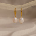 A close-up view of the Dangling Pearl Drop Earrings delicately hanging from a textured beige ribbon. Each earring features a luminous, oval-shaped freshwater pearl suspended from a sleek, high-polish gold lever-back hook. The soft lighting highlights the pearlescent sheen and natural texture of the pearls, while the neutral backdrop enhances the earrings’ timeless elegance and minimal design.