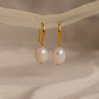A close-up view of the Dangling Pearl Drop Earrings delicately hanging from a textured beige ribbon. Each earring features a luminous, oval-shaped freshwater pearl suspended from a sleek, high-polish gold lever-back hook. The soft lighting highlights the pearlescent sheen and natural texture of the pearls, while the neutral backdrop enhances the earrings’ timeless elegance and minimal design.
